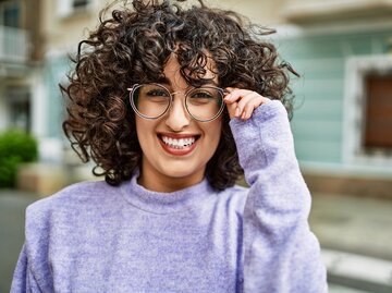 Lächelnde Frau mit braunen Locken und Brille vor bunter Hausfassade | © AdobeStock/Krakenimages.com