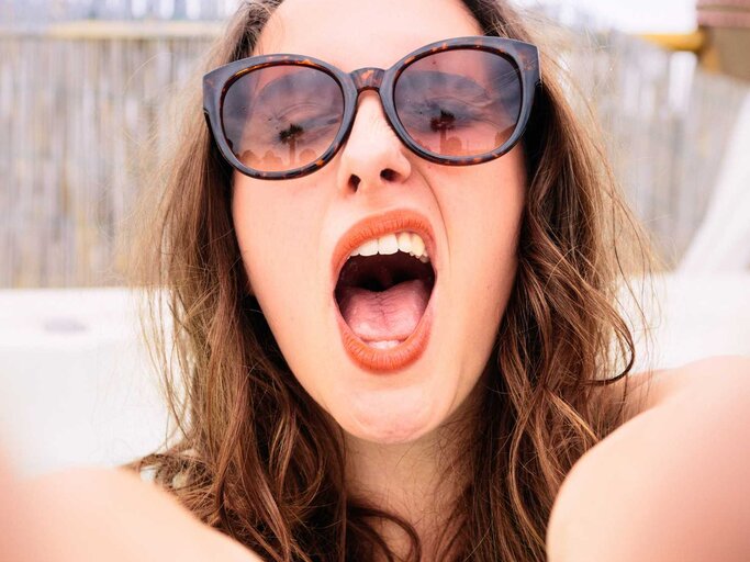 Junge Frau mit Sonnenbrille macht frech ein Selfie. | © Getty Images/georgeclerk
