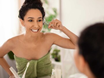 Frau benutzt Wimpernzange vor dem Spiegel und lächelt | © Getty Images/Prostock-Studio