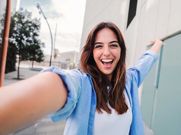 Frau steht vor einem Gebäude und macht freudestrahlend ein Selfie. | © Adobe Stock/Jose Calsina