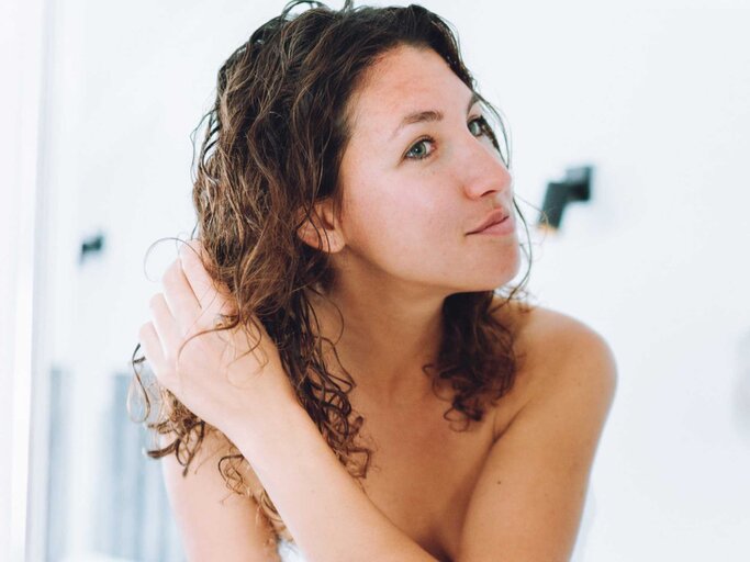 Frau steht mit nassen Haaren im Bad und schaut sich ihre Haare im Spiegel an | © Getty Images/Olga Rolenko