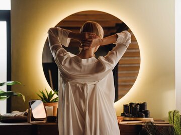 Frau steht im Morgenmantel vor dem Spiegel und greift mit den Händen ihre Haare am Hinterkopf zusammen. | © Adobe Stock/Drobot Dean