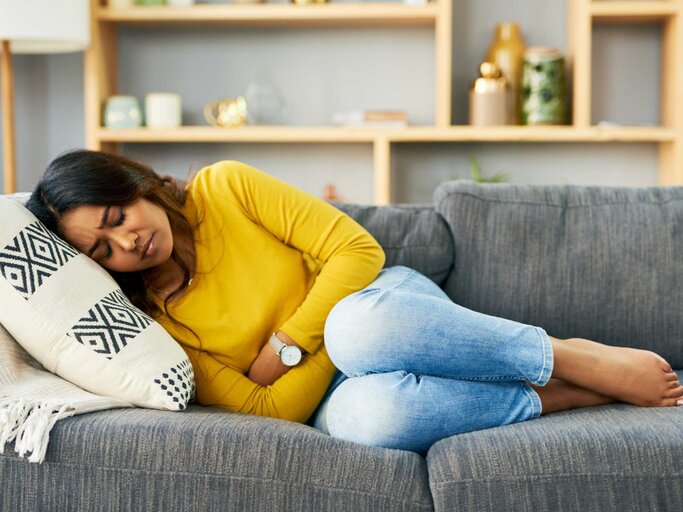 Frau liegt mit Menstruationsschmerzen auf der Couch | © Getty Images/Moyo Studio