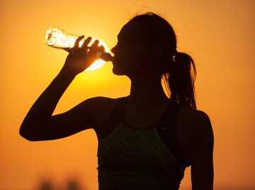 Frau mit Pferdeschwanz steht im Sonnenuntergang und trinkt aus einer Plastikflasche | © AdobeStock/V&P Photo Studio
