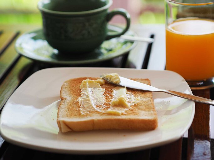 Auf einem Frühstückstisch mit Orangensaft und Kaffee liegt ein Toast mit Butter auf einem Teller. | © Getty Images / Penpak Ngamsathain