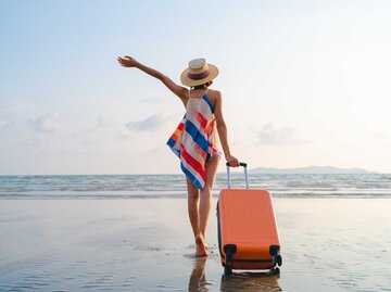 Frau steht mit ihrem Koffer am Strand und hebt euphorisch den Arm | © Getty Images/Twenty47studio