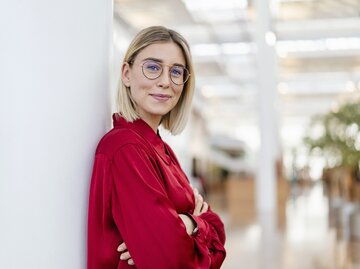 Junge blonde Frau trägt Brille, rote Bluse und lächelt in die Kamera. Ihre Arme sind verschränkt und sie lehnt sich gegen eine Wand | © Getty Images/Westend61