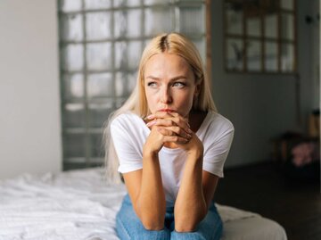 Porträt einer depressiven Frau, die allein zu Hause sitzt und mit traurigem Gesichtsausdruck wegschaut, Hände am Kinn haltend | © Getty Images/dikushin