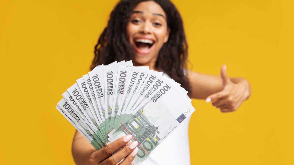Aufgeregte afroamerikanische Frau hält mehrere Hunderteuro Scheine in der Hand und zeigt mit dem Finger auf Banknoten, sie steht vor einem gelbem Hintergrund. | © Adobe Stock/Prostock-studio