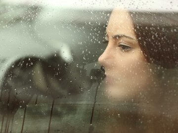 Eine junge Frau mit braunen Haaren sitzt im Auto und schaut aus dem Fenster, welches von Regentropfen bedeckt ist. | © Adobe Stock/Antonioguillem