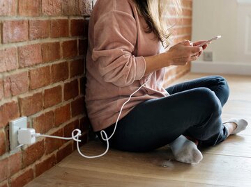 Eine Frau sitzt an einer Backsteinwand und benutzt ein Smartphone, das an ein Ladekabel angeschlossen ist. | © Getty Images/Rawpixel.com