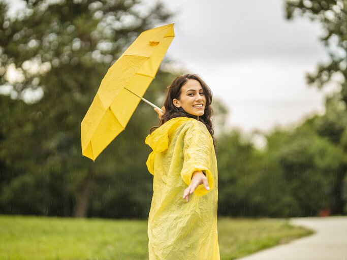 Eine Frau im Regenmantel und mit Regenschirm streckt die Hand nach hinten aus | © GettyImages/Eleganza