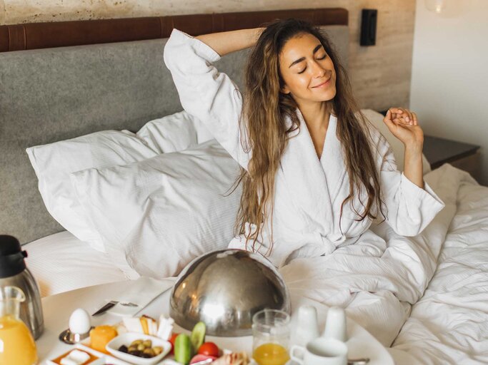 Frau sitzt im Bett im Hotelzimmer und frühstückt. | © Getty Images / Oleg Breslavtsev
