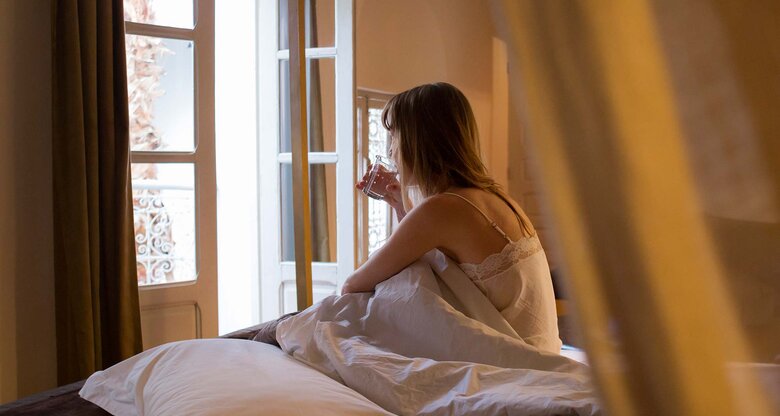 Frau sitzt mit Nachthemd im Bett und schaut mit einem Glas Wasser aus dem geöffneten Fenster. | © Getty Images/Kathrin Ziegler