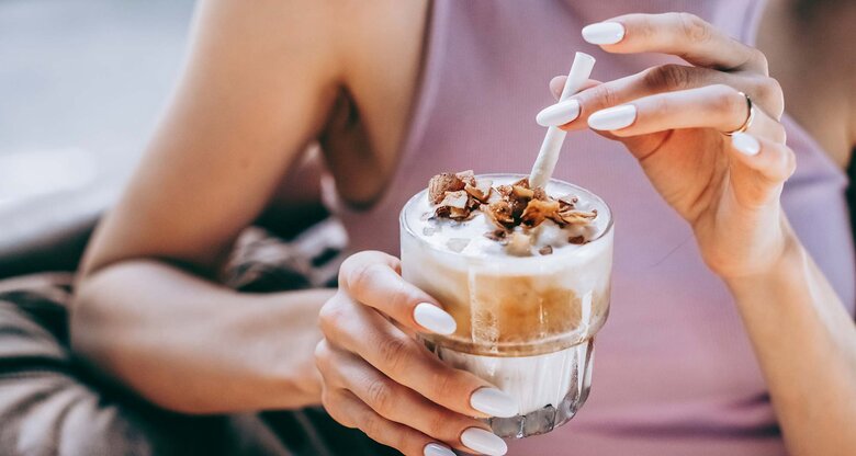 In einem lilafarbenen Top hält eine Frau ein Glas Eiskaffee mit Milchschaum und Schokostückchen, bereit zum ersten Schluck. | © Getty Images/Milky Way