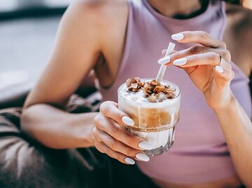 In einem lilafarbenen Top hält eine Frau ein Glas Eiskaffee mit Milchschaum und Schokostückchen, bereit zum ersten Schluck. | © Getty Images/Milky Way