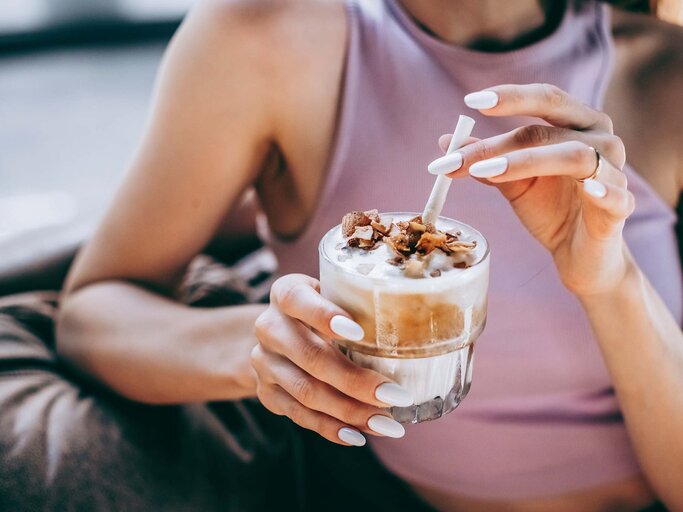 In einem lilafarbenen Top hält eine Frau ein Glas Eiskaffee mit Milchschaum und Schokostückchen, bereit zum ersten Schluck. | © Getty Images/Milky Way