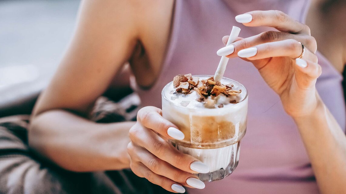 In einem lilafarbenen Top hält eine Frau ein Glas Eiskaffee mit Milchschaum und Schokostückchen, bereit zum ersten Schluck. | © Getty Images/Milky Way