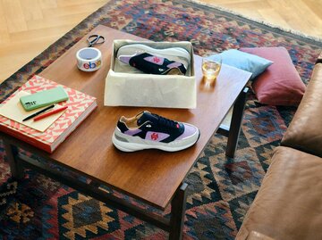 Wohnzimmer mit brauner Ledercouch und braunem Tisch auf gemustertem Teppich, darauf verschiedene Produkte wie Sneaker platziert. | © eBay.de