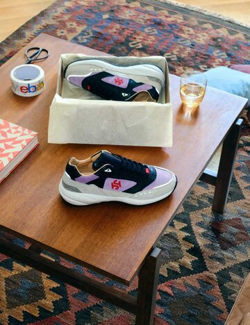 Wohnzimmer mit brauner Ledercouch und braunem Tisch auf gemustertem Teppich, darauf verschiedene Produkte wie Sneaker platziert. | © eBay.de