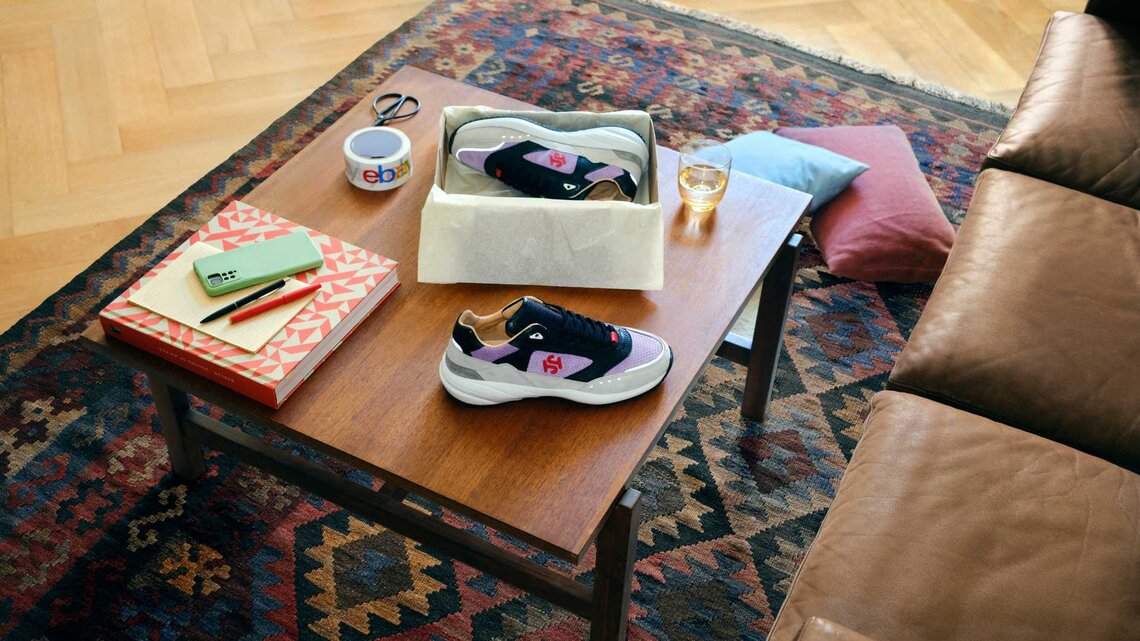 Wohnzimmer mit brauner Ledercouch und braunem Tisch auf gemustertem Teppich, darauf verschiedene Produkte wie Sneaker platziert. | © eBay.de