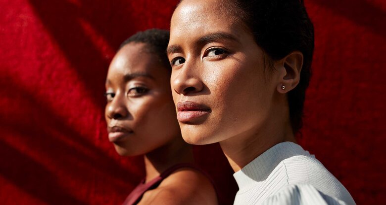 Zwei junge Frauen mit ernstem Blick stehen im Sonnenlicht vor einem roten Hintergrund, eine in Weiß, die andere in Bordeauxrot gekleidet. | © Getty Images/Klaus Vedfelt
