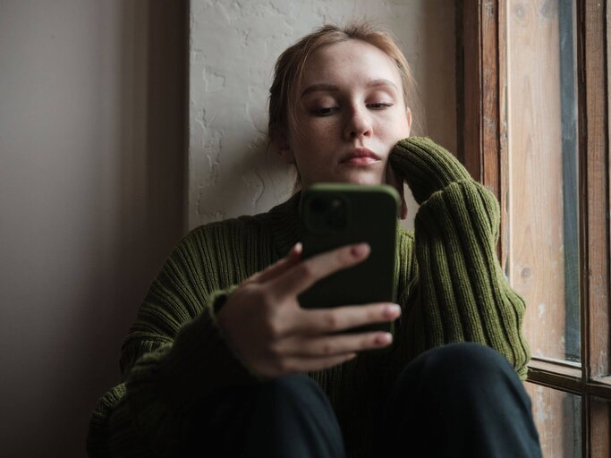 Person sitzt an Fenster mit Handy in der Hand | © Getty Images/MementoJpeg