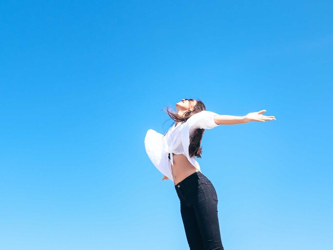 Glückliche Frau, die frische Luft atmet, die Arme in die Höhe hebt und einen sonnigen blauem Himmel genießt | © Adobe Stock/max_play