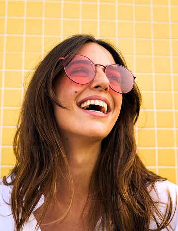 Brünette mit Sonnenbrille auf lacht und blickt nach oben | © Getty Images/Carlos Barquero