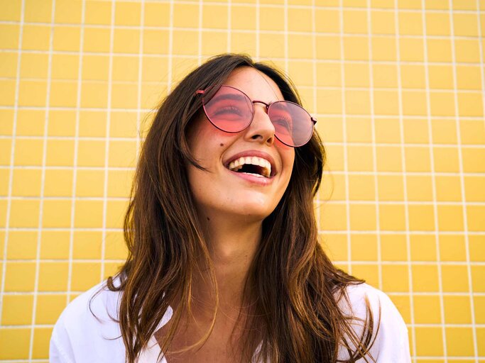 Brünette mit Sonnenbrille auf lacht und blickt nach oben | © Getty Images/Carlos Barquero