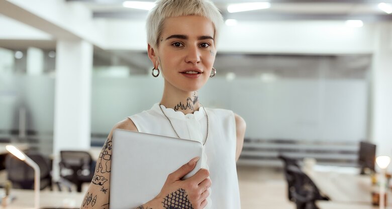 Junge Frau mit kurzen, blonden Haaren und Tattoos steht selbstbewusst in einem Büro | © AdobeStock/Svitlana