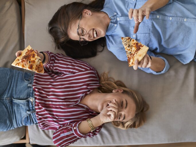 Zwei Freundinnen liegen lachend auf der Couch und essen Pizza | © 	GettyImages/Westend61