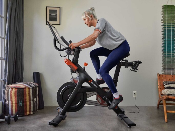 Frau trainiert zuhause auf einem Peloton Bike, trägt Sportkleidung und AirPods, während sie auf den integrierten Bildschirm schaut. Im Hintergrund ist ein gemütlich eingerichteter Raum mit Sessel, Teppich und Wanddeko zu sehen. | © Peloton