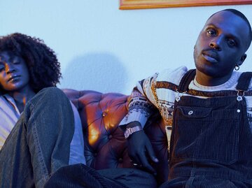 Junger Mann mit Latzhose sitzt entspannt im kühlen Licht auf einem Sofa und schaut intensiv in die Kamera, neben ihm eine Frau mit Locken. | © Getty Images/Delmaine Donson
