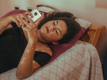 Frau mit vielen Sommersprossen liegt im Bett und scrollt durch ihr Handy | © GettyImages/isabelladias