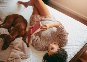 Frau liegt mit ihrem Hund lesend im Bett