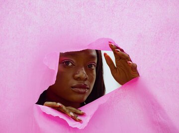 Afroamerikanische Frau blickt durch einen Riss in einer pinken Wand | © GettyImages/TatsianaVolkava