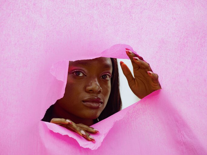 Afroamerikanische Frau blickt durch einen Riss in einer pinken Wand | © GettyImages/TatsianaVolkava