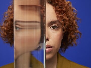 Junge Frau mit roten Locken blickt ernst in die Kamera, ihr Gesicht halb hinter einem Glas mit verzerrtem Spiegelbild verborgen. | © GettyImages/Klaus Vedfelt