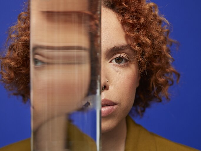 Junge Frau mit roten Locken blickt ernst in die Kamera, ihr Gesicht halb hinter einem Glas mit verzerrtem Spiegelbild verborgen. | © GettyImages/Klaus Vedfelt