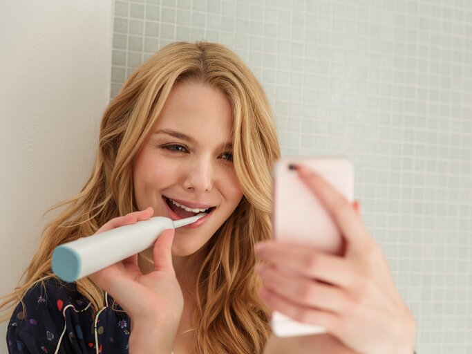 Eine junge Frau putzt sich die Zähne und ist am Handy | © GettyImages/Lilly Bloom