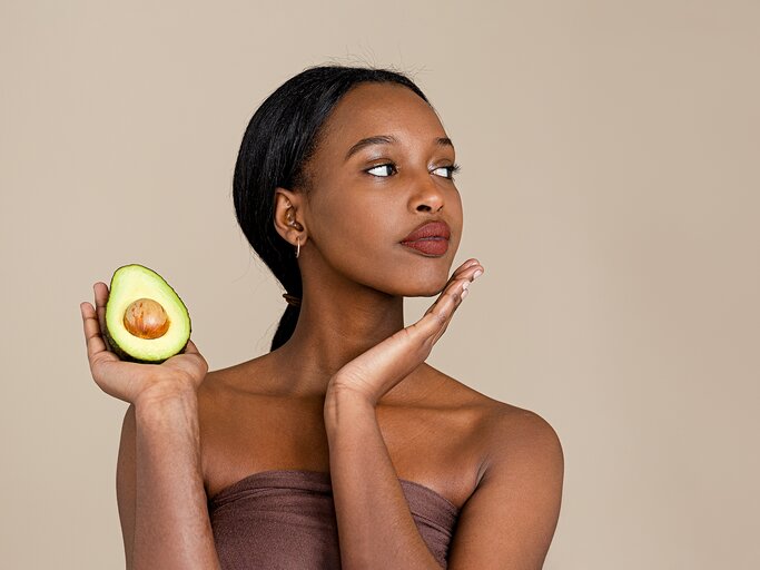 Eine junge Frau mit schöner Haut hält eine halbe Avocado neben ihr Gesicht | © GettyImages/Phamai Techaphan