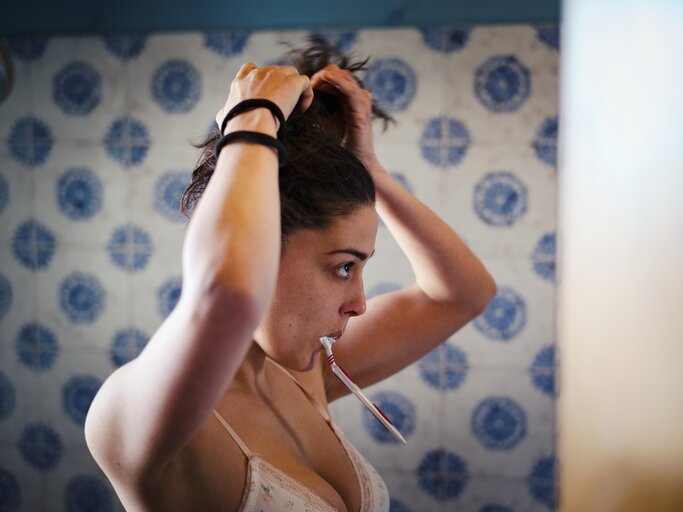 Frau macht sich vor dem Spiegel einen Dutt, während sie ihre Zähne putzt | © Getty Images/Justin Case