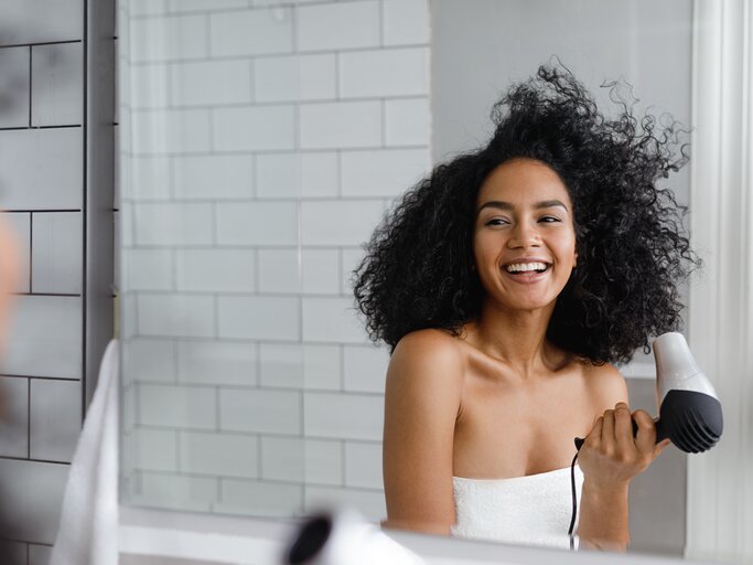 Frau föhnt sich die Haare und betrachtet sich lächelnd im Spiegel | © Getty Images/Artem Varnitsin / EyeEm