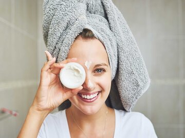 Junge Frau hält sich Gesichtscreme vor das Auge und lächelt | © Getty Images/Moyo Studio