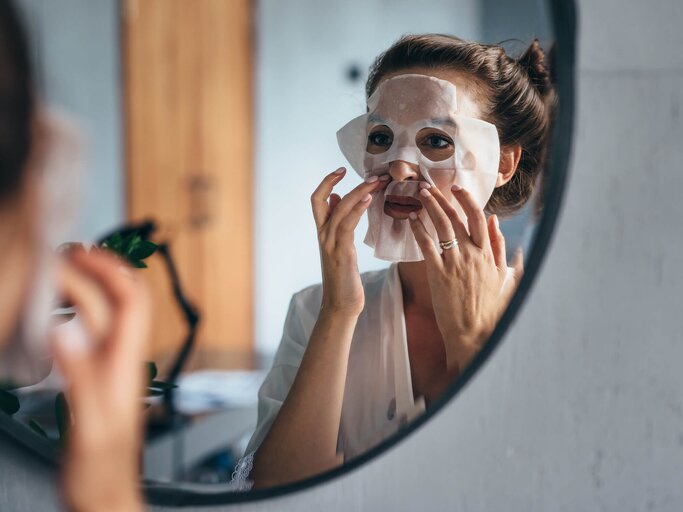 Frau legt vor dem Spiegel eine Tuchmaske auf ihr Gesicht | © Getty Images/undrey