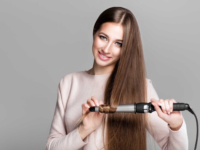 Schöne lächelnde Frau, die lange Haare mit Lockenstab lockt. | © Getty Images/Erstudiostok