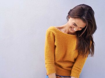 Frau mit orangenem Pullover und langen braunen Haaren steht vor einer Wand und lächelt fröhlich. | © Getty Images / Yuri_Arcurs