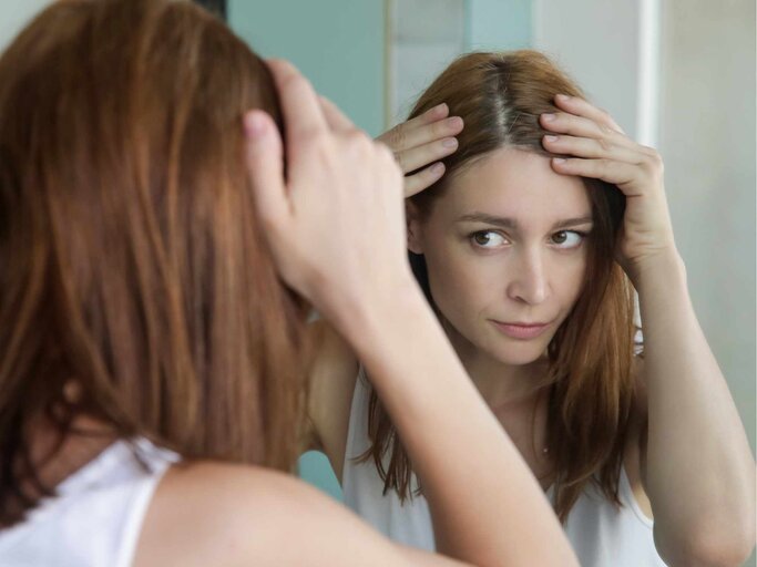 Porträt einer schönen jungen Frau, die ihre Kopfhaut und Haare vor dem Spiegel, Haarwurzeln, Farbe, grauehaare, Haarausfall oder trockene Kopfhaut Problem untersucht | © Getty Images/triocean