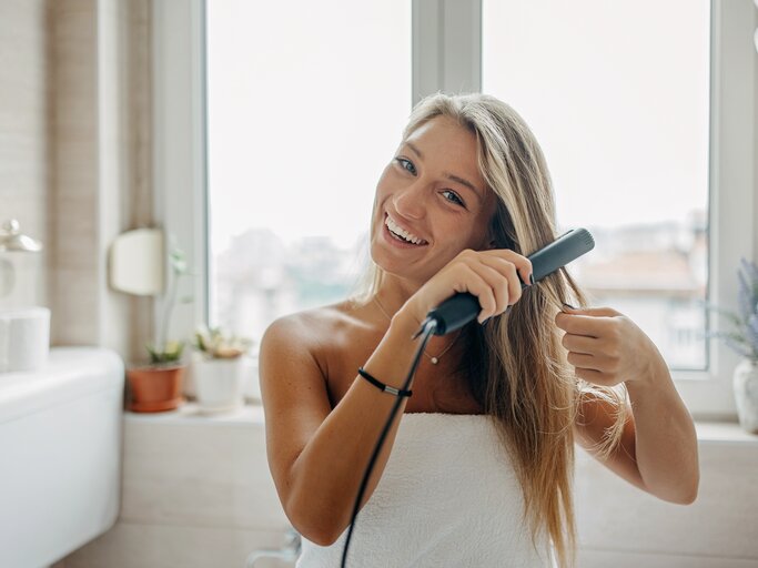 Junge blonde Frau lächelt während sie sich die Haare glättet | © Getty Images/supersizer